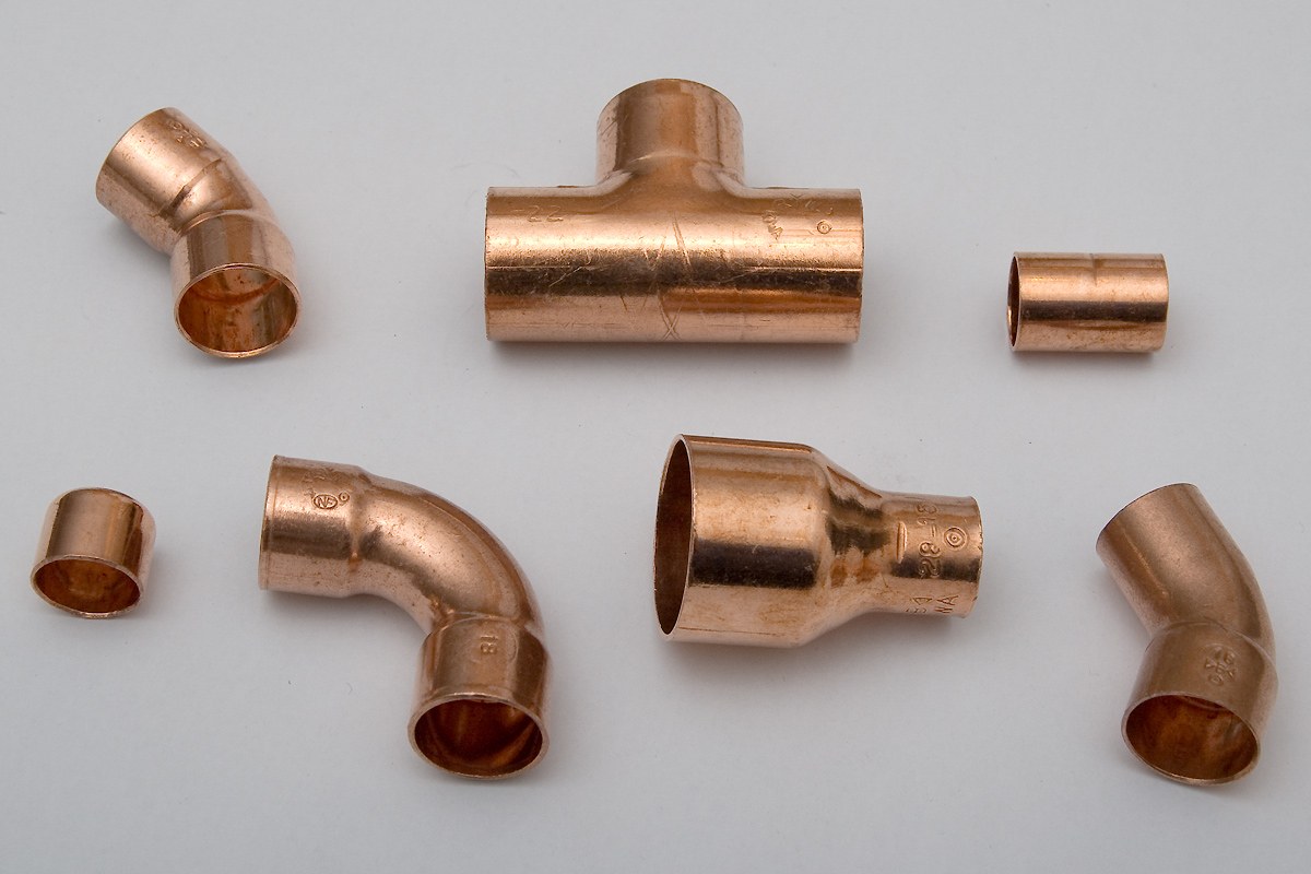 Seven copper pipe fittings laid out on a grey surface: elbows, a tee, couplings and a reducer