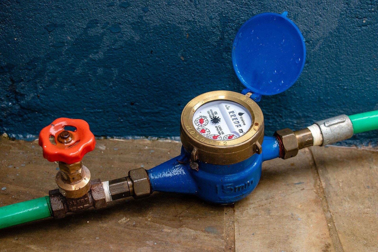 A blue water meter and a red-wheeled gate valve with green pipe tails, resting on a bare floor