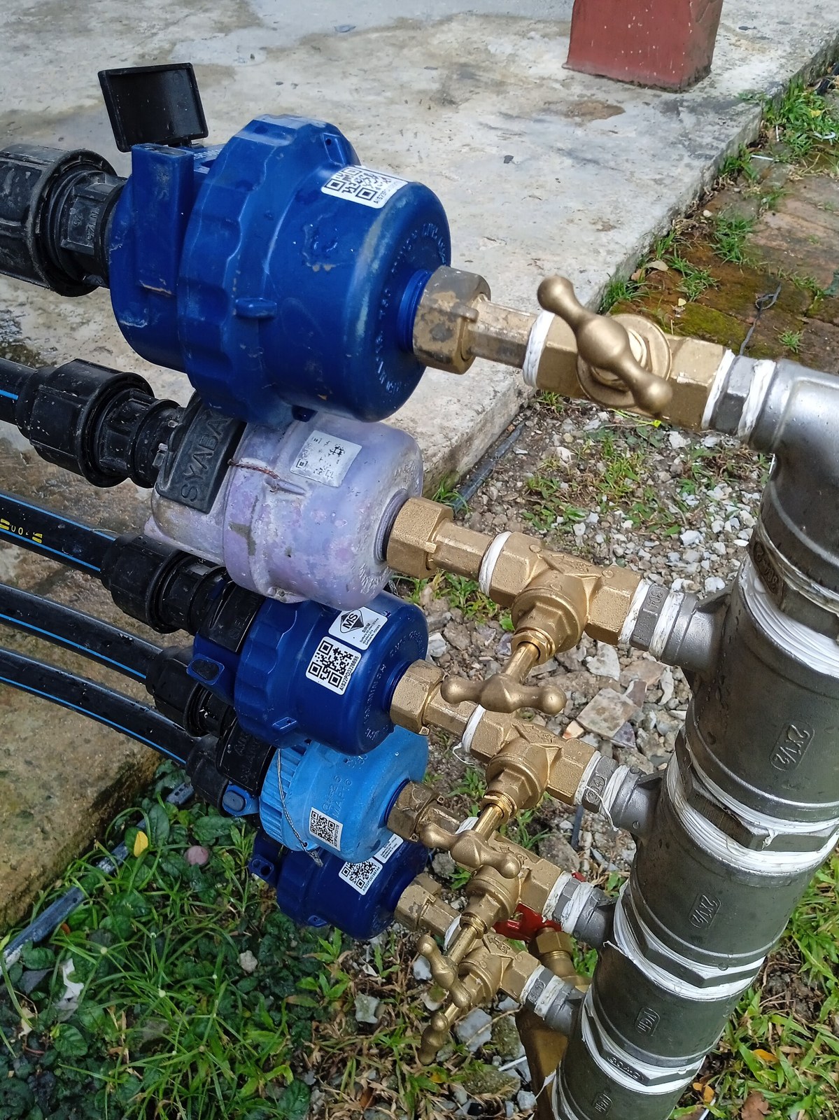 A bank of four water meters and brass valves standing at ground level beside a kerb