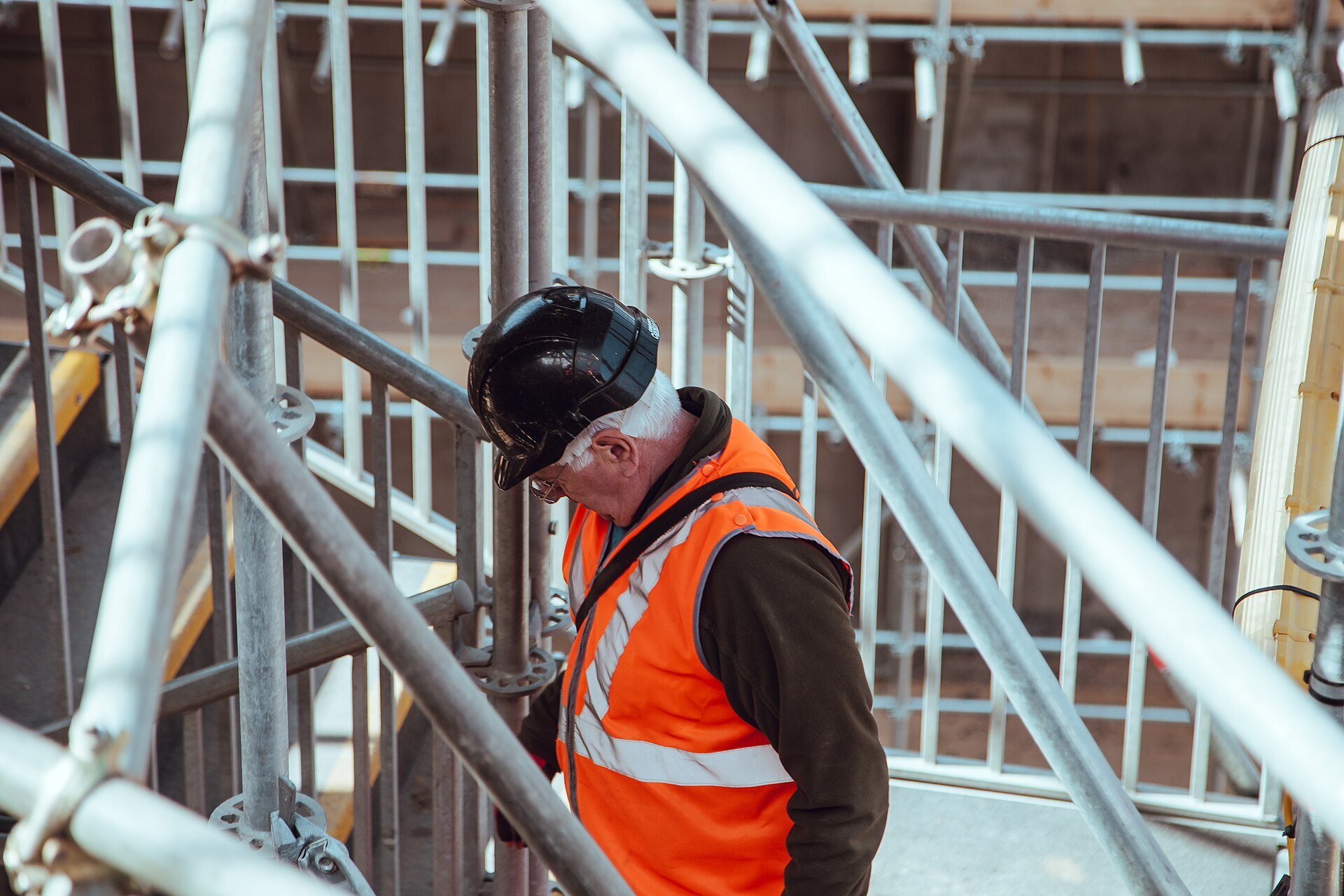 A worker in a black hard hat and orange high-visibility vest climbing scaffold stairs.