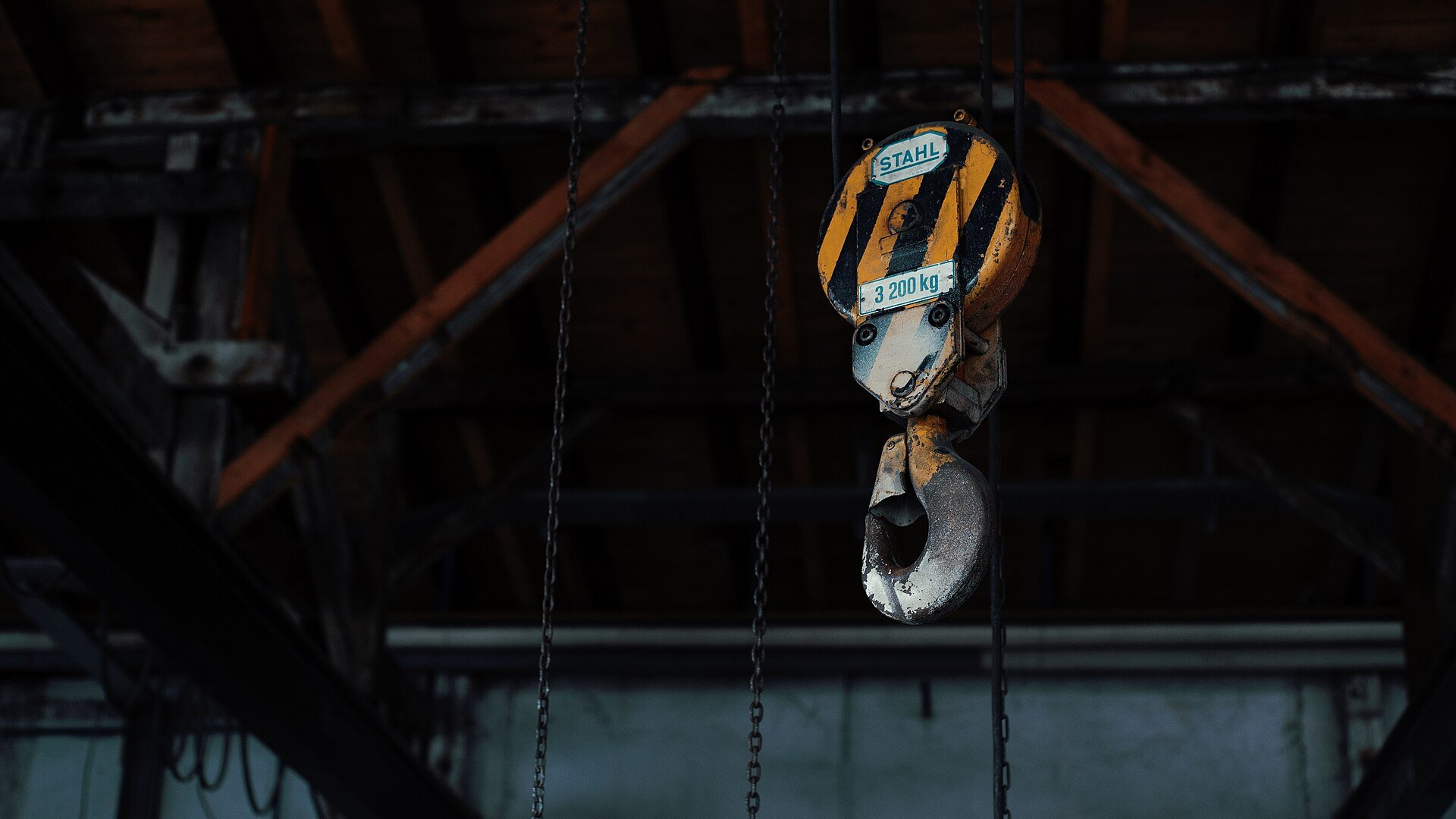 A yellow and black crane hook block hanging from chains under a dark steel roof.