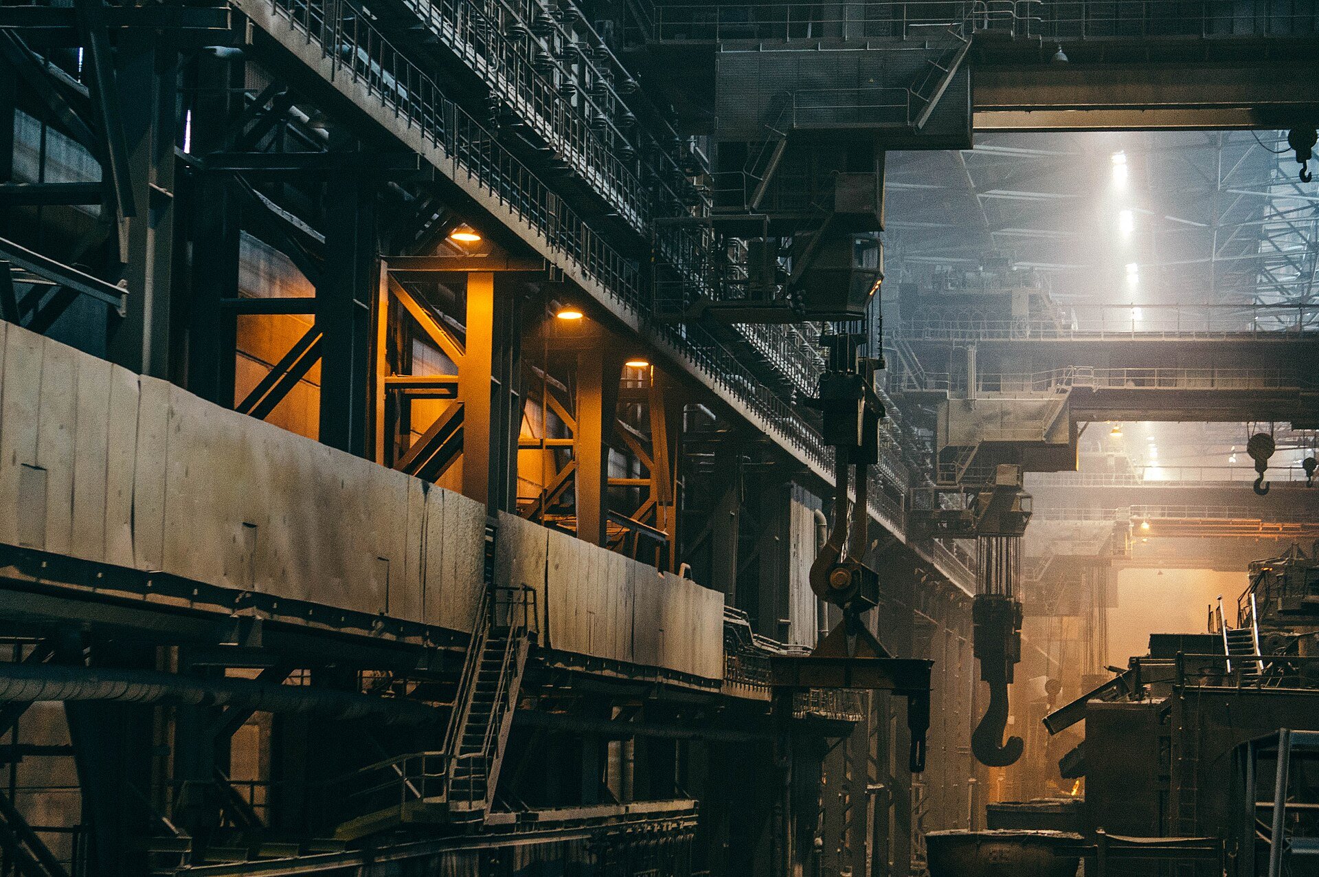 The interior of a steel plant with overhead cranes, hooks and walkways lit in amber.