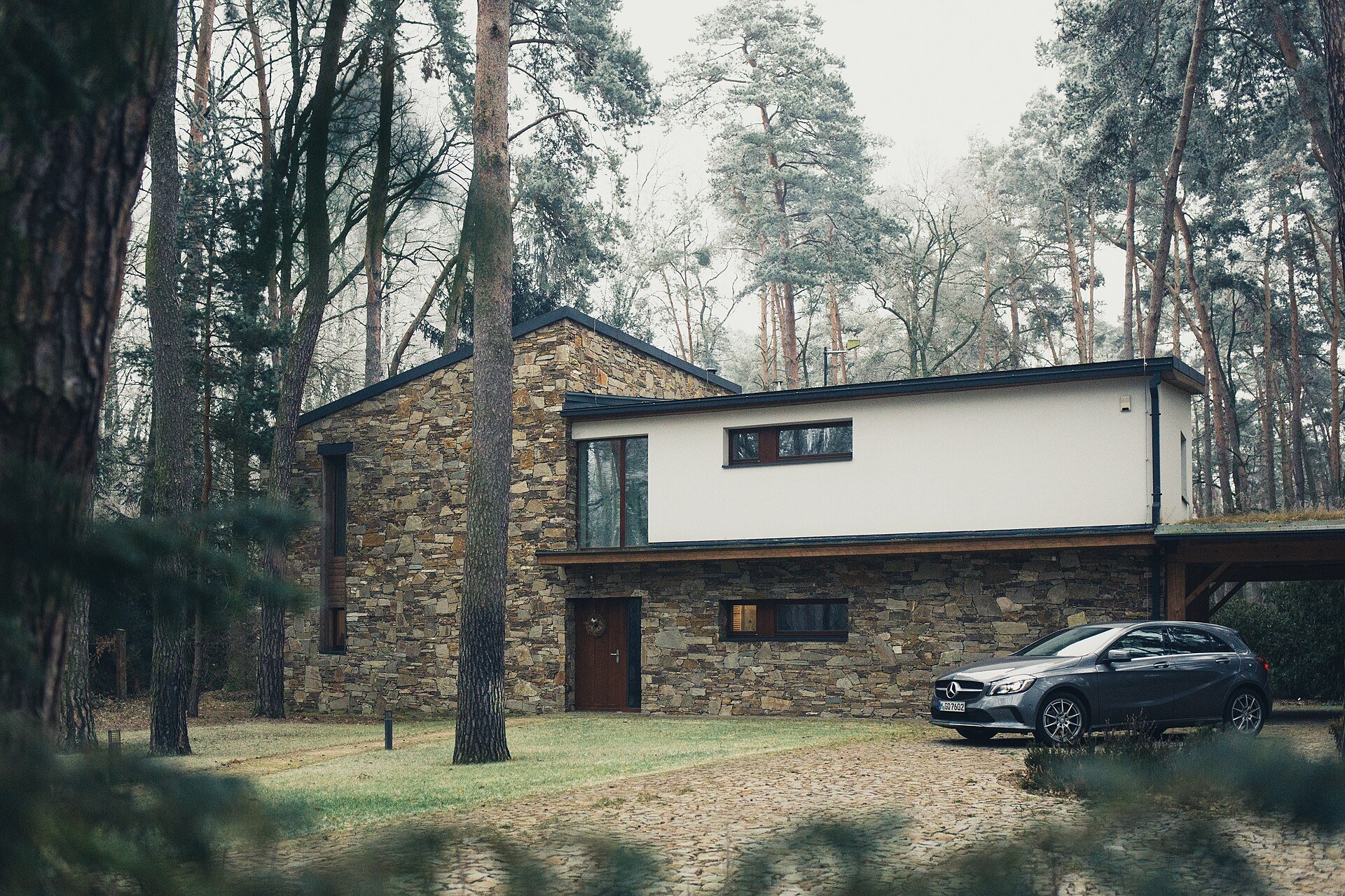 A stone and white-rendered house among tall pines, with a grey car in the driveway.