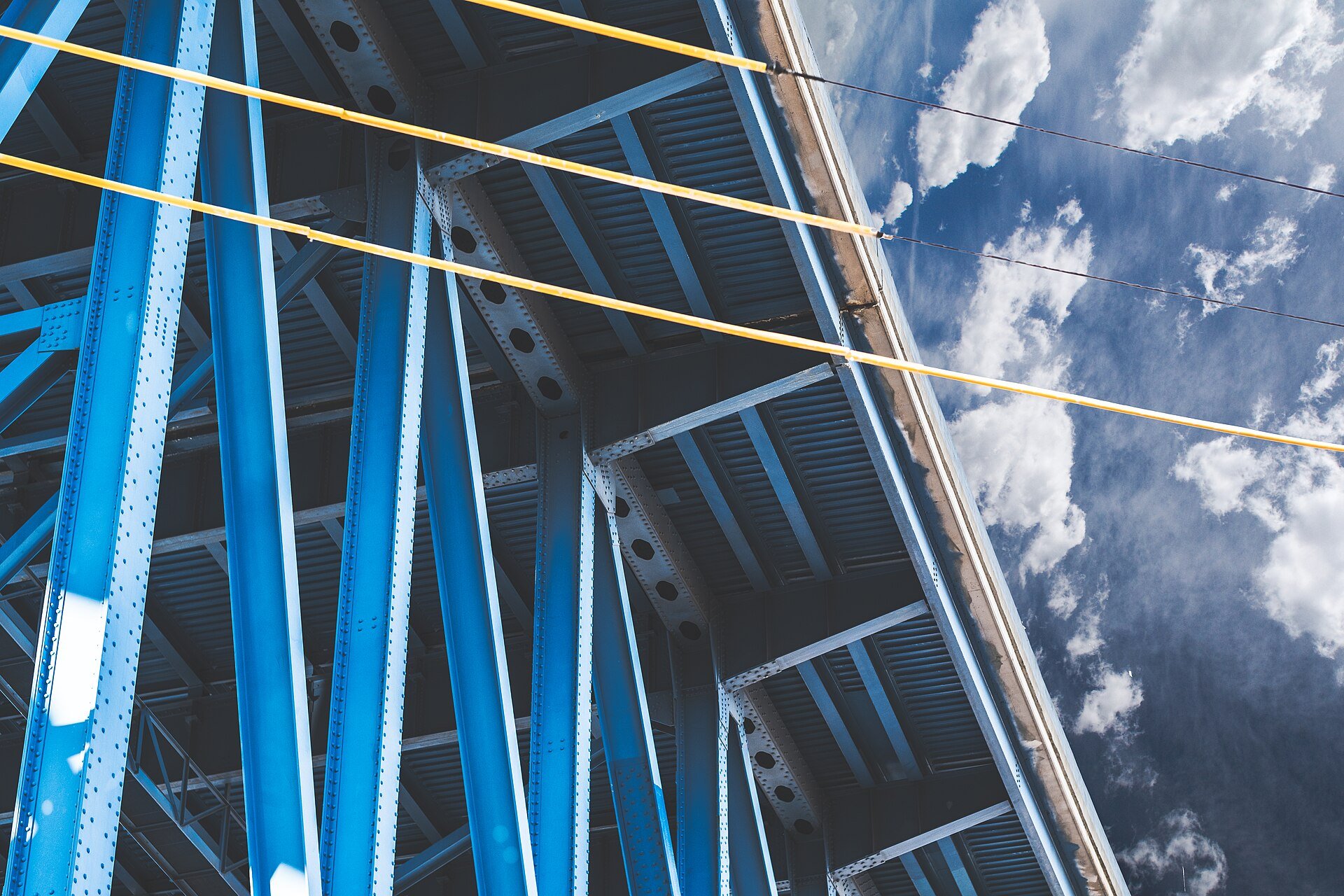 Blue riveted steel girders seen from below against a cloudy sky, crossed by yellow cables.
