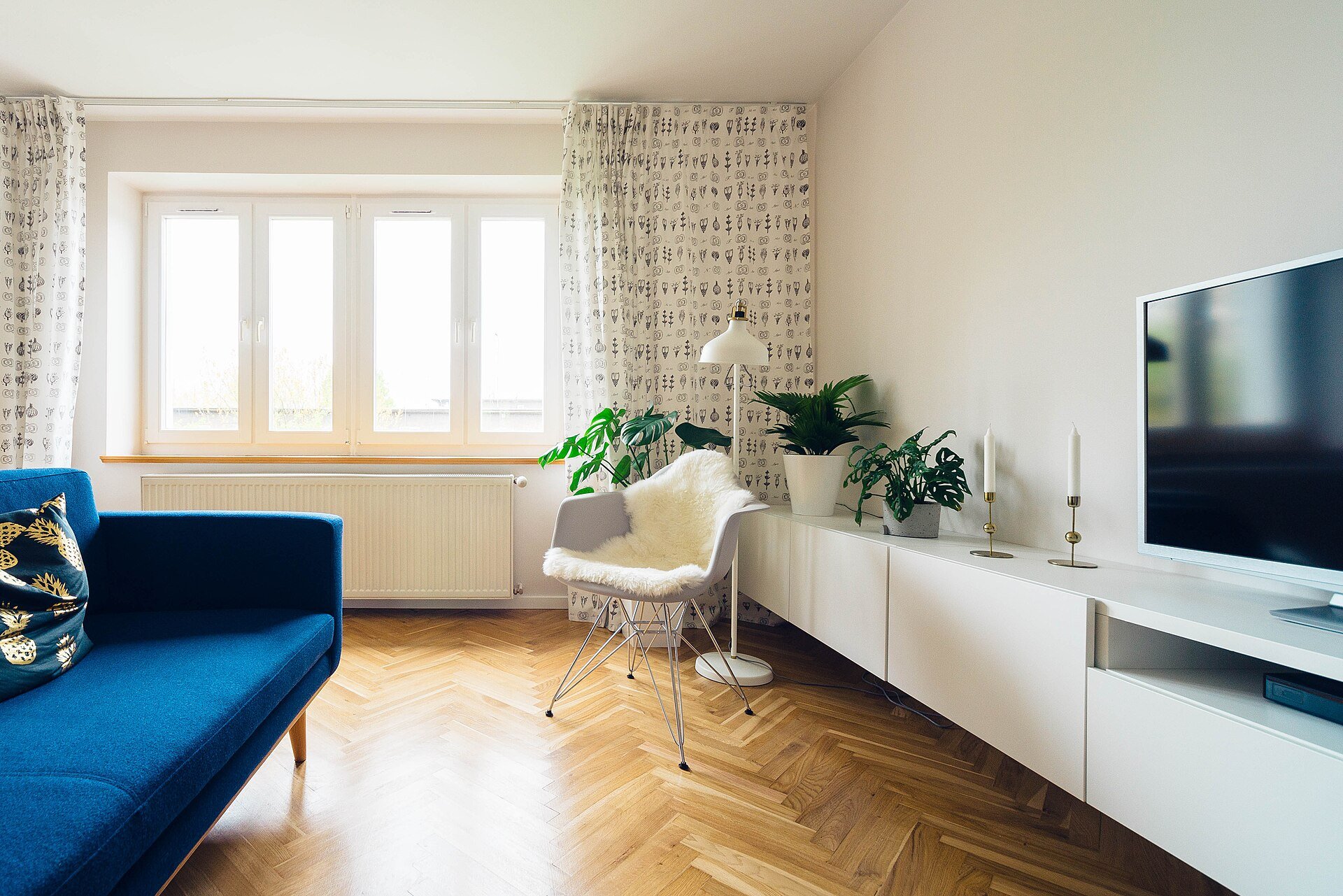 A bright living room with a blue sofa, a white chair, a floor lamp and a television.