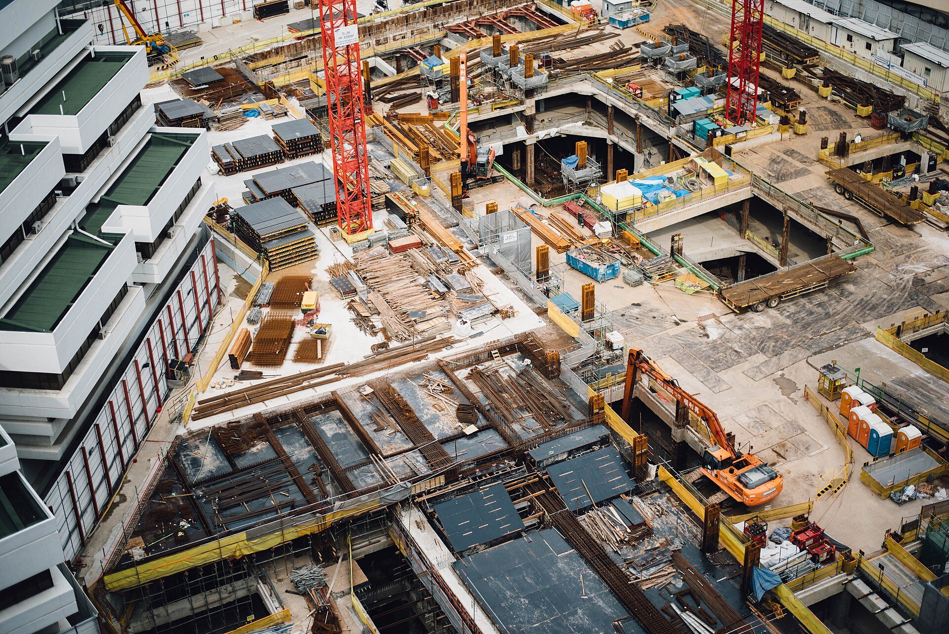 An aerial view of a large construction site with red tower cranes, rebar and an excavator.